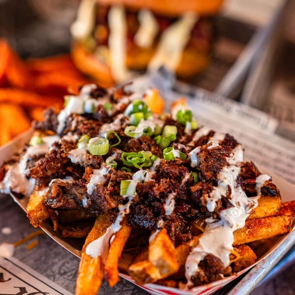 Loaded fries with meat, white sauce, and green onions, with a burger in the background.