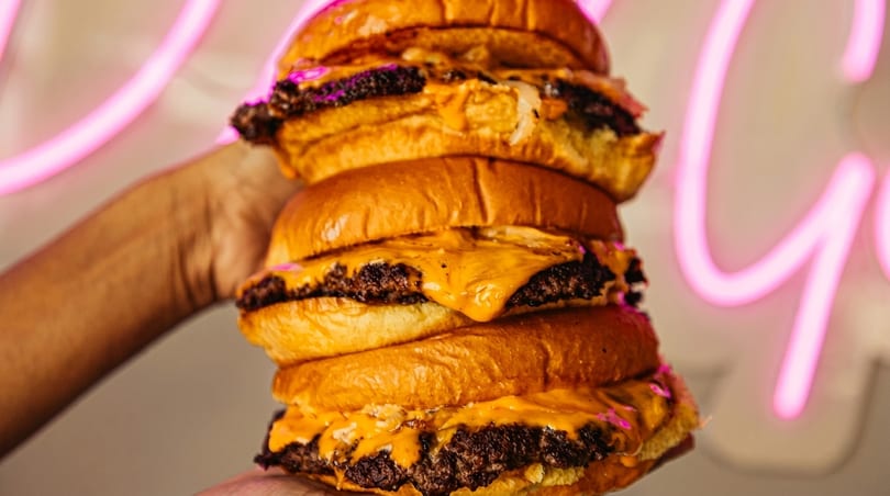 A hand holds a towering stack of four cheeseburgers with melted cheese and beef patties.