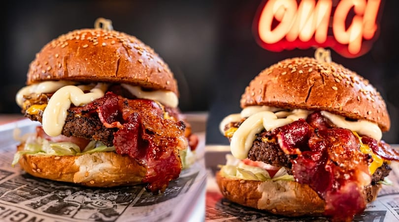 Two juicy bacon cheeseburgers with mayonnaise and lettuce on a newspaper-lined tray.