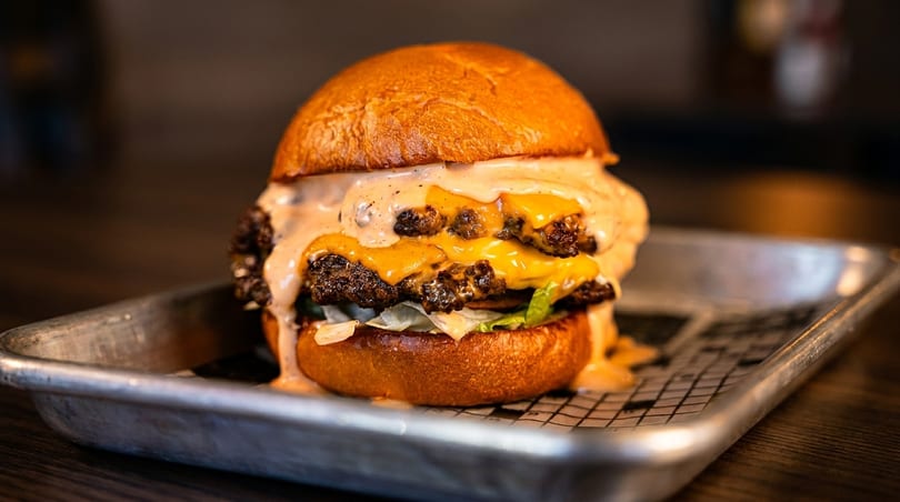 A double cheeseburger with melted cheese, special sauce, and lettuce on a brioche bun, served on a metal tray.