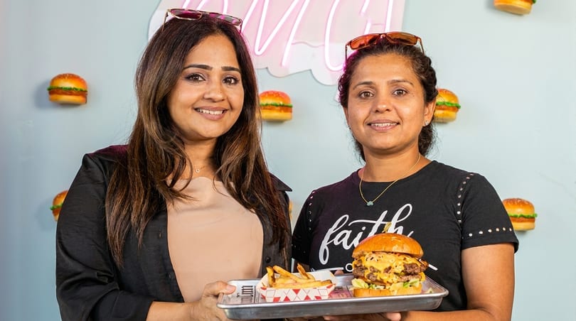 Two women smiling, holding a tray with a double cheeseburger and fries against a burger-themed wall.