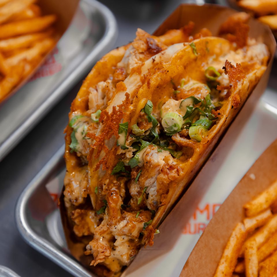 A crispy chicken taco with sauce and green onions, served with fries on a metal tray.