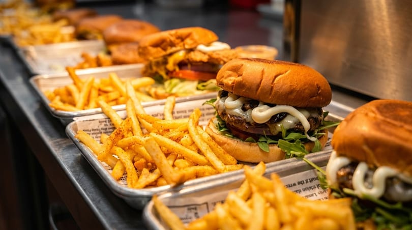 Multiple trays of burgers with melted cheese, arugula, and fries line a counter.
