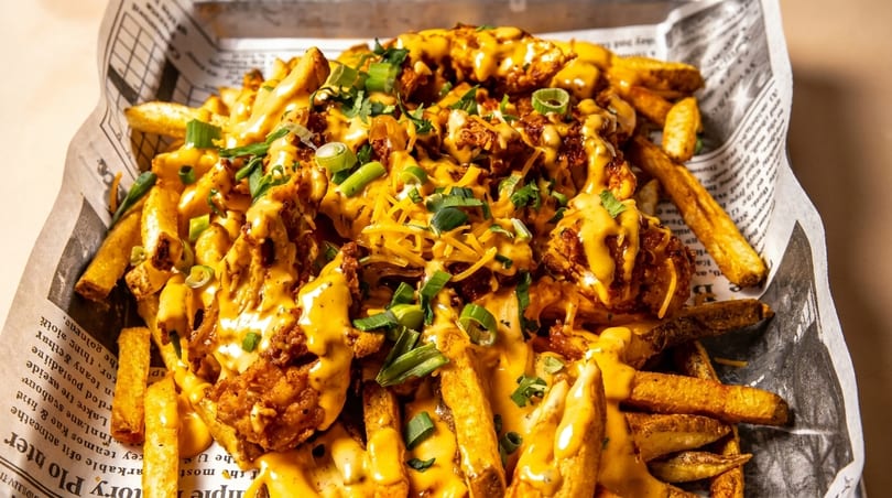 Crispy fries topped with melted cheese, chicken, shredded cheese, and green onions, served in a newspaper-lined basket.