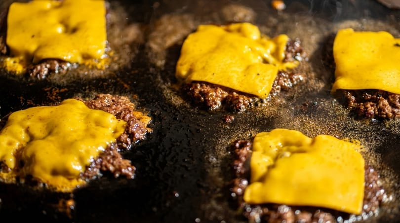 Close-up of several smashburger patties with melting yellow cheese cooking on a hot griddle.