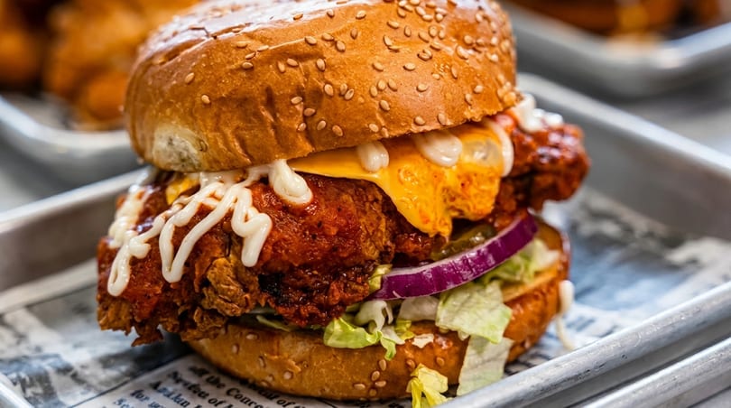 Spicy fried chicken sandwich with melted cheese, red onion, lettuce, and white sauce on a sesame bun.