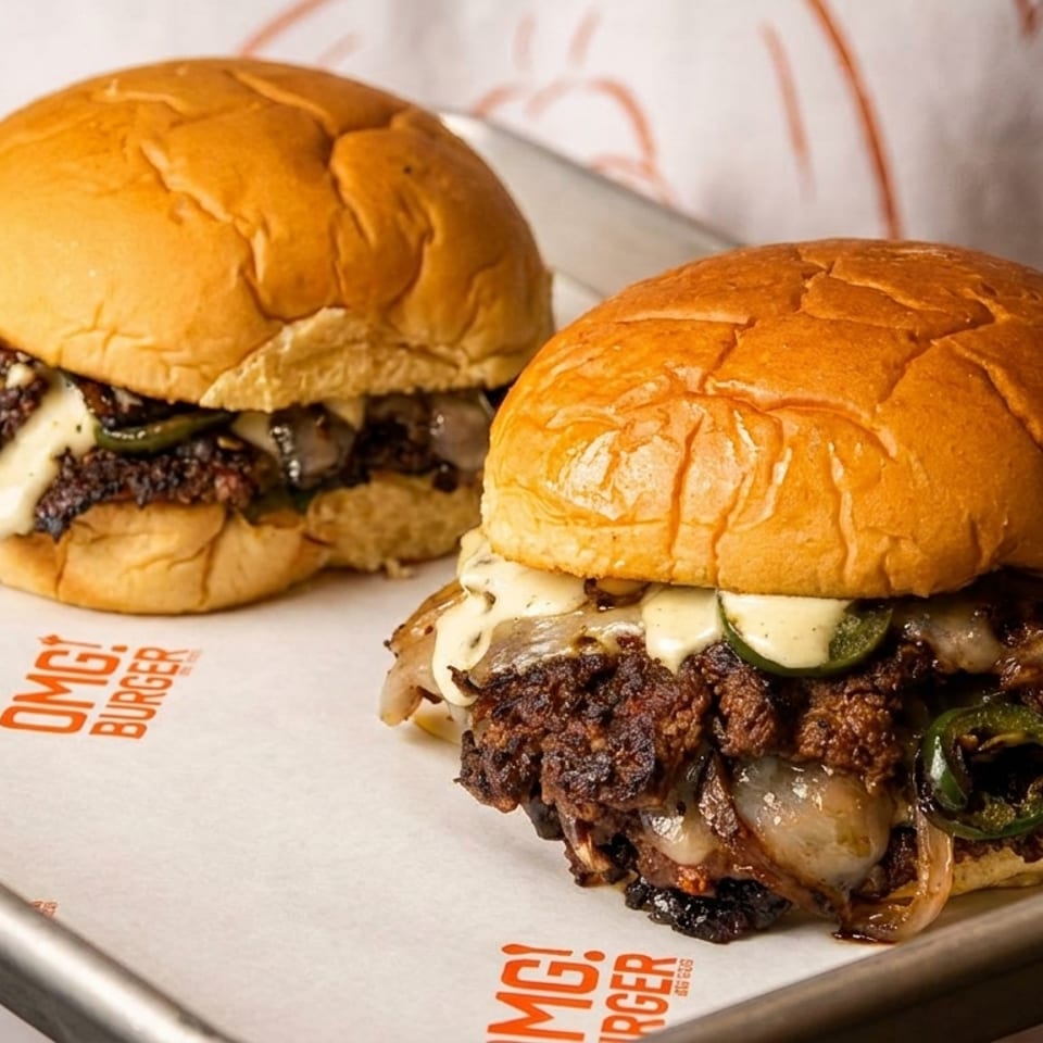 Two delicious OMG! Burgers with melted cheese, grilled onions, and jalapeños on a metal tray.